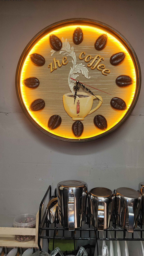 ساعت دیواری چوبی با طراحی خاص برای کافه ژ، شامل چیدمان دانههای قهوه به جای اعداد ساعت، تصویر فنجانی با بخار در حال بالا رفتن در مرکز، و عناصر گرافیکی مینیمال با رنگهای گرم؛ القاکنندهی فضایی دنج و صمیمی