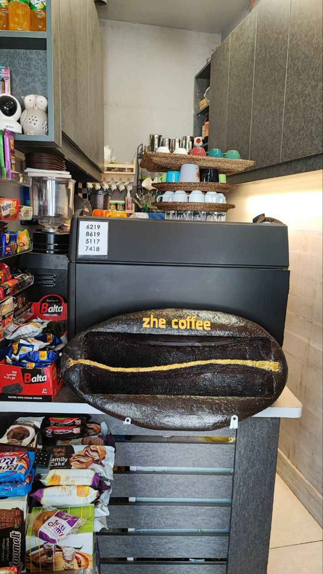 ظرف خوراکی به شکل دانهی قهوه با نوشتهی «zhe coffee» در محیط کافه، همراه با خوراکیهای بستهبندیشده و عناصر مکمل مانند قهوهساز و تزئینات دیواری مرتبط با تم قهوه؛ مناسب برای استفاده در دکوراسیون کافه یا سرو تنقلات