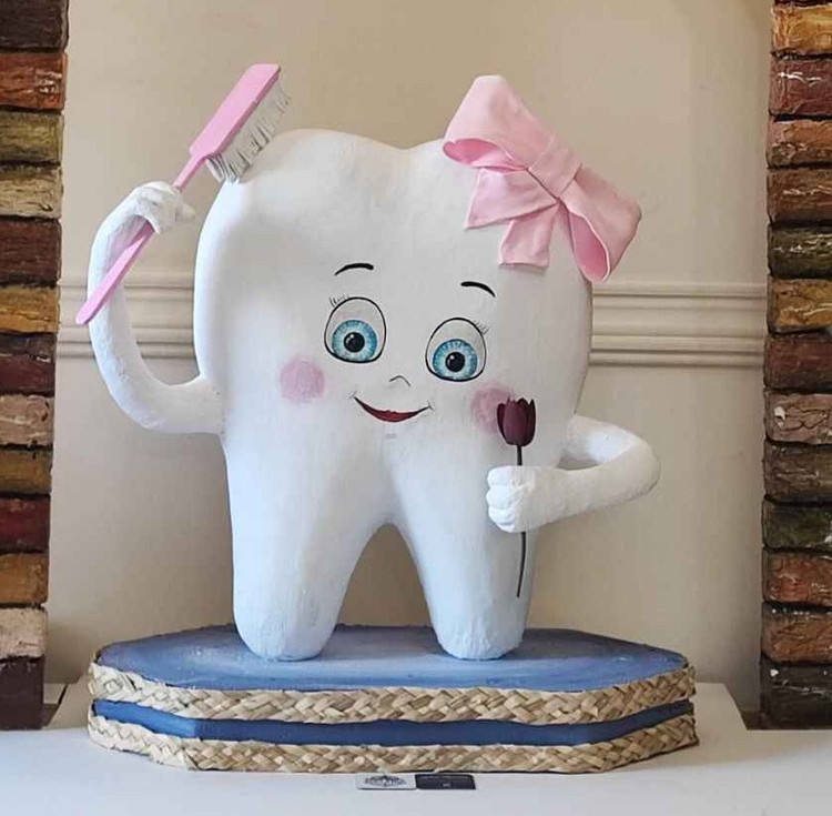ماکت شخصیت دندان با چشمان آبی، لبخند کودکانه، پاپیون صورتی، همراه با مسواک و گل تزئینی؛ قرارگرفته روی پایه آبی با تزئین حصیری، مناسب فضاهای کلینیکی و کودکمحور